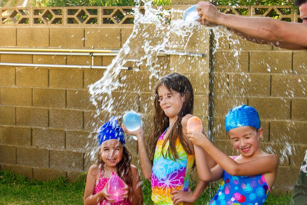 The arms of a father break a blue water balloon over the heads of his three daughters. The girls are wearing bathing suits and two of them have swim caps on.