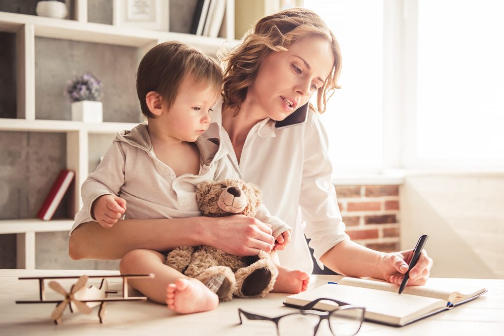 Beautiful business mom is talking on the mobile phone and taking notes while spending time with her cute baby boy at home