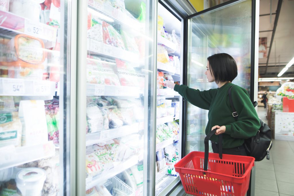 A girl with a basket takes frozen foods from the supermarket's refrigerator. The girl chooses goods in the store. Shopping in the supermarket