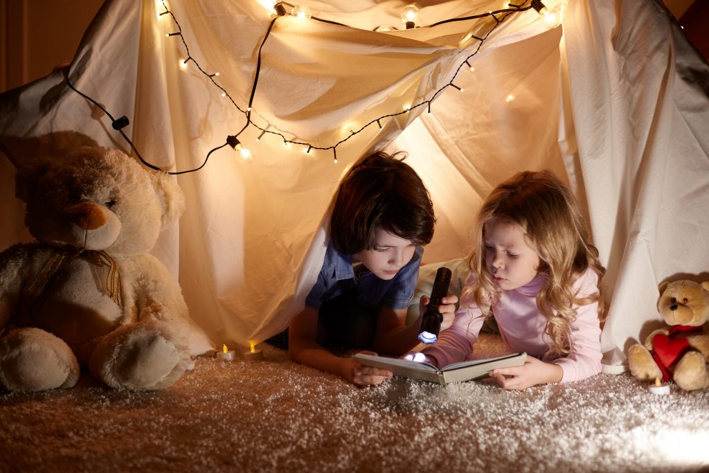 Little kids involving in reading amazing book. They lying in nice toy tent in playroom. Boy holding flashlight in hand