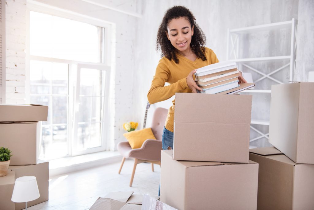 Favorite books. Pleasant curly-haired girl packing a pile of books into a big box before moving out of the room in the dormitory