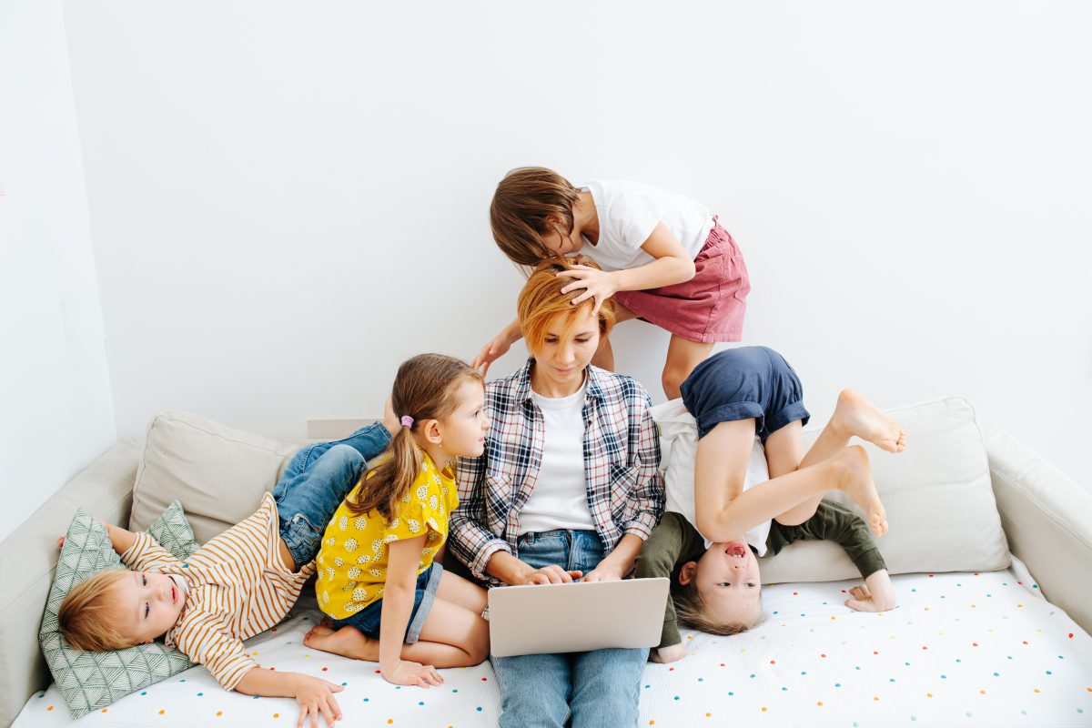 Young mom working on a laptop on a sofa in the living room, while her wild kids literally play on top of her head. They jump, roll and make noises. Tips and Tricks for Boosting Productivity as a Stay-at-Home Mom