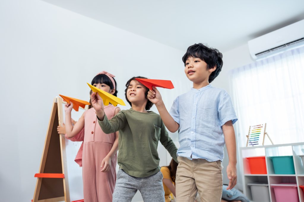 Group of Mixed race young little kid playing airplane in schoolroom. Child girls and boys student spending time play paper rocket while during break with feeling fun together in classroom at school.