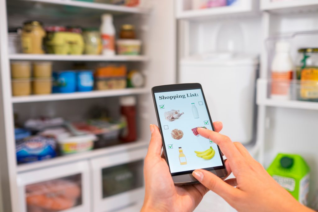 Woman makes her shopping list on his phone connected to the refrigerator